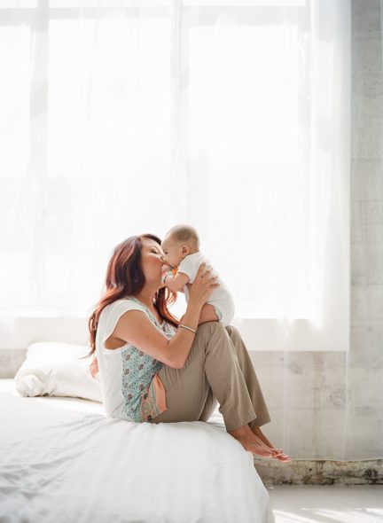 Asian mom sitting on white bed kissing baby boy