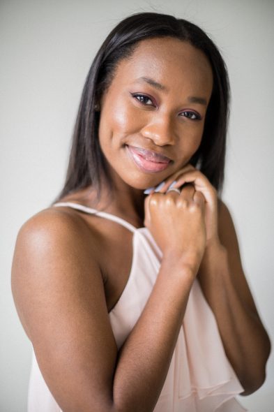 young, black woman in light pink sleeveless top smiling at camera with hands clasped together under her chin, dimples